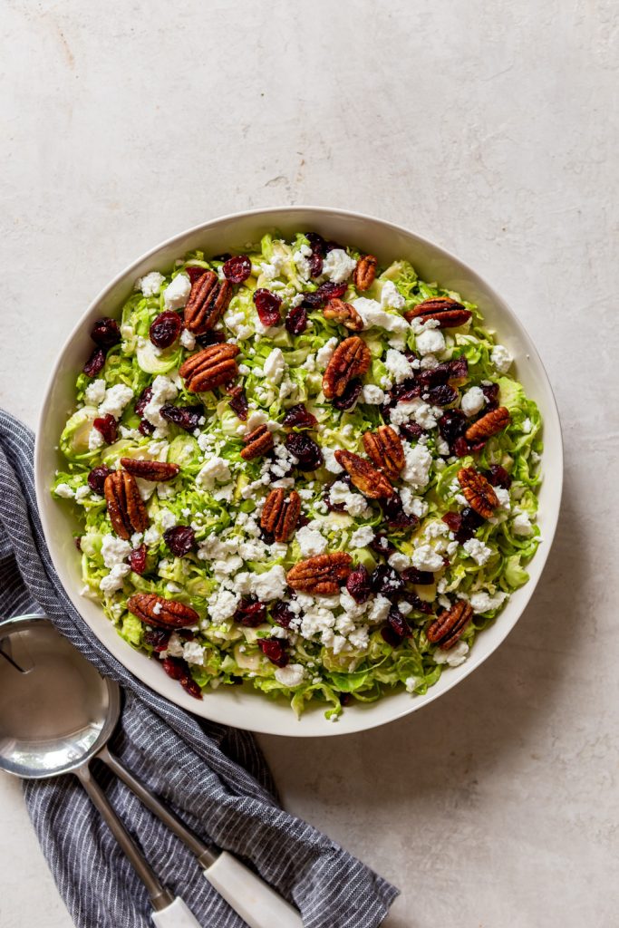 Ensalada de coles de Bruselas afeitada con queso de cabra, arándanos secos y nueces confitadas, en un bol junto a un paño de cocina azul.