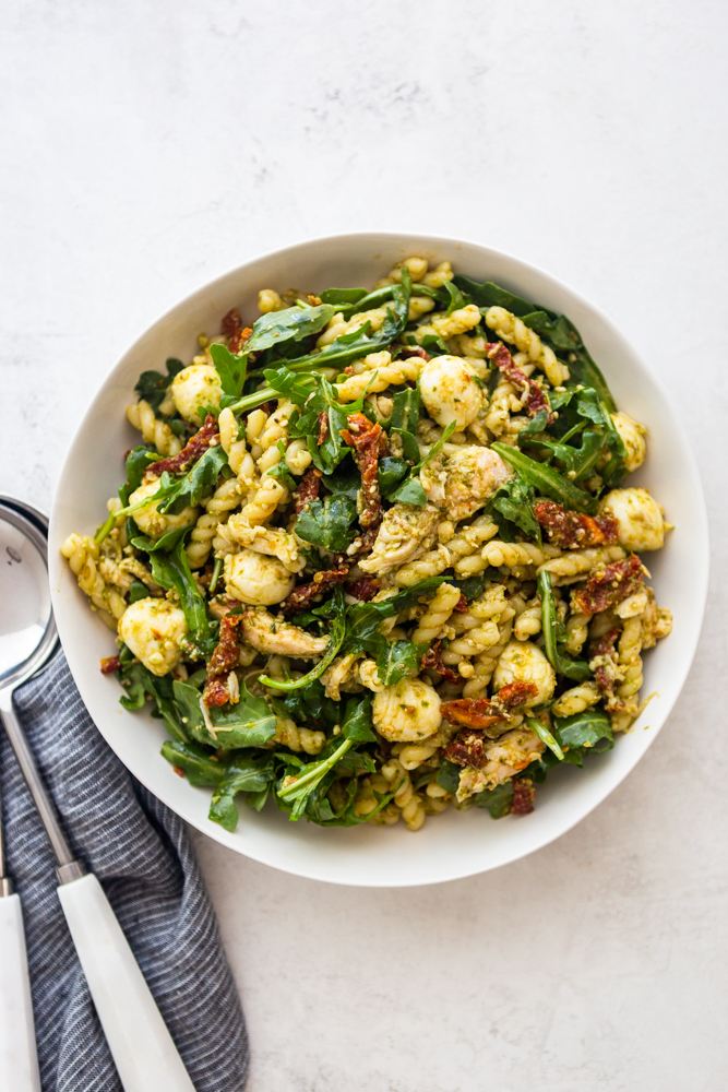 Ensalada de pasta con pesto de pollo en un tazón blanco con un paño de cocina a rayas azules y una cuchara para servir