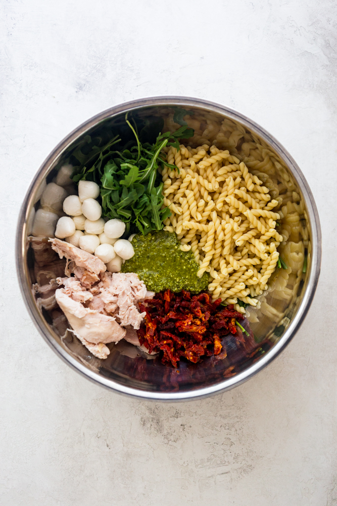 Ingredientes para Ensalada de pasta con pollo al pesto: pasta hervida, rúcula, bolas de mozzarella, pollo hervido, tomates secados al sol, pesto, todos juntos en un tazón grande de acero inoxidable