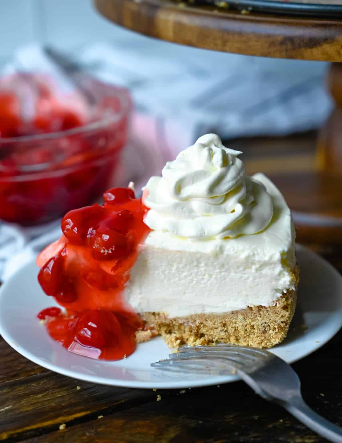 Un trozo de tarta de queso en un pequeño plato blanco con crema batida y cerezas encima.
