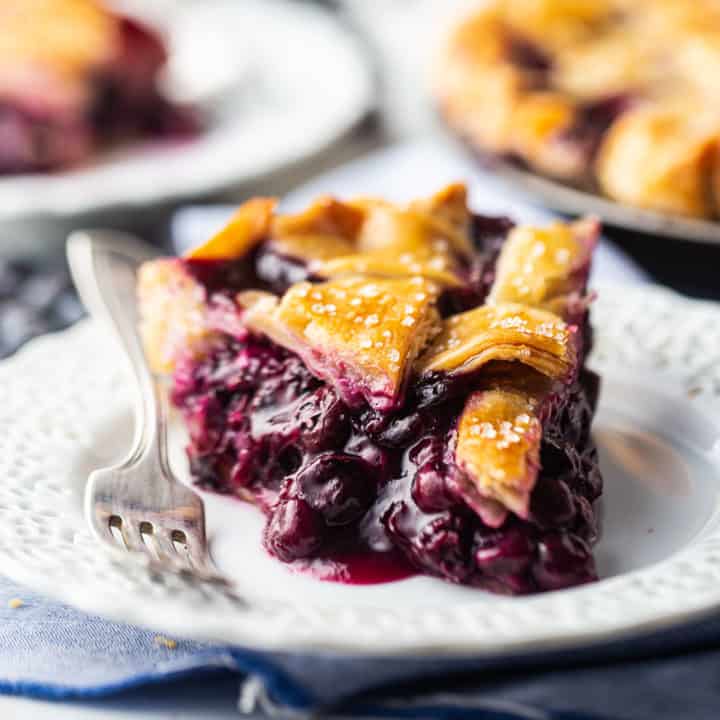 Una rebanada de pastel de arándanos con relleno grueso y corteza superior enrejada, presentada en un plato de encaje blanco.