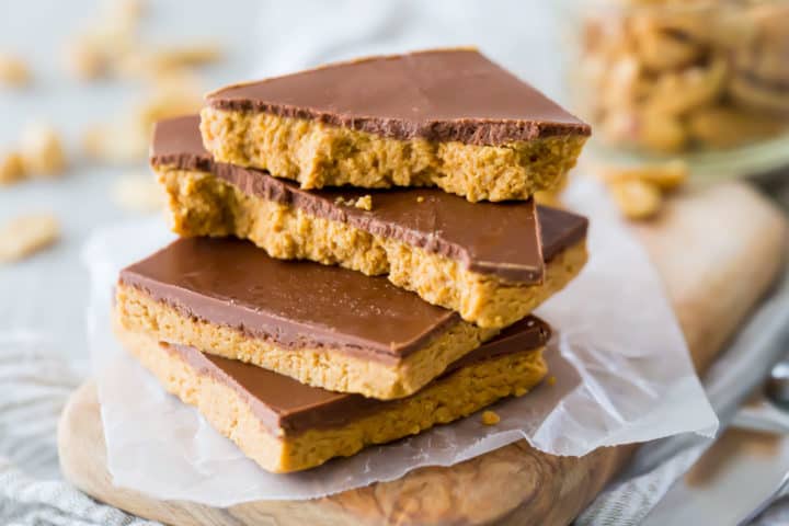 Barras de chocolate con mantequilla de maní apiladas con un frasco de maní en el fondo.