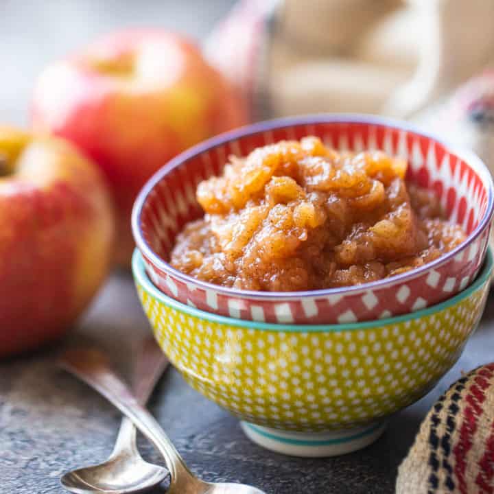 Salsa de manzana en un recipiente colorido, con manzanas al fondo.