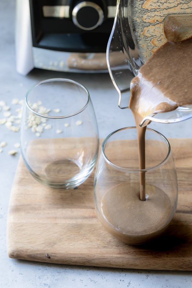 Verter el batido del desayuno en un vaso vacío con otro vaso al lado y la máquina Vitamix de fondo