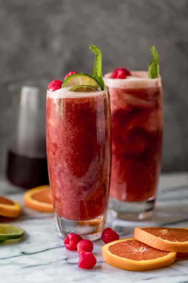 Dos vasos altos de batido de té helado de frambuesa con rodajas de naranja, gajos de limón, frambuesas frescas y hojas de menta adornadas en la bebida. Jarra de té helado o frambuesa en el fondo