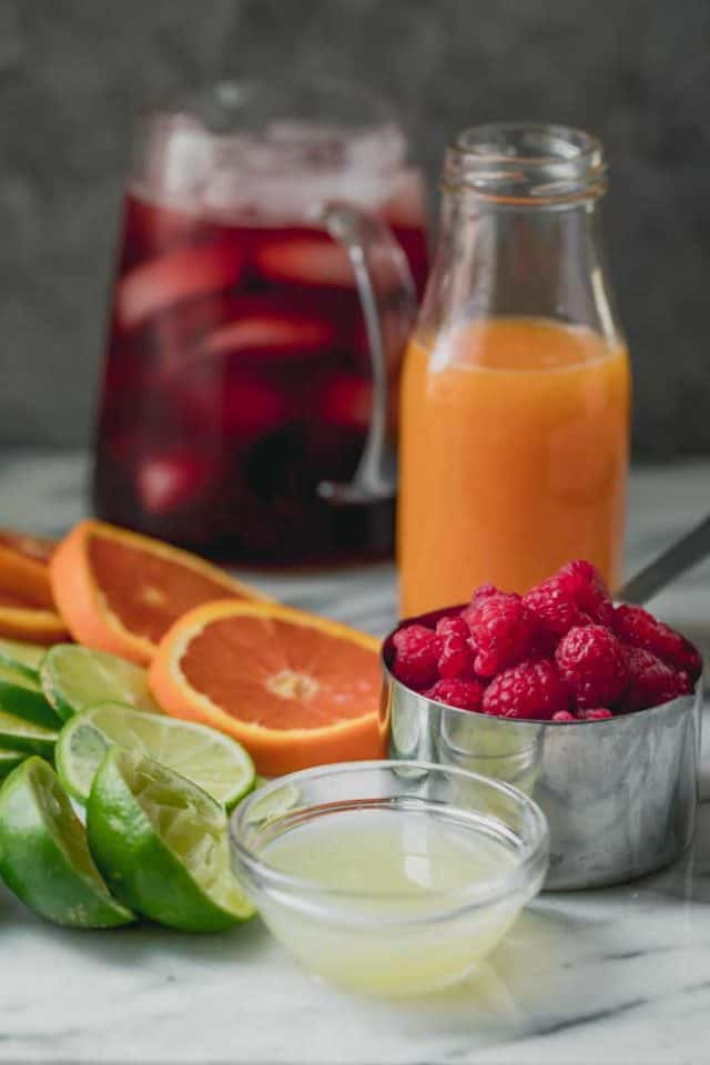 Ingredientes listos para el batido de té helado de frambuesa - Jarra de té helado de frambuesa con cubitos de hielo, vaso de jugo de naranja, tazón de jugo de limón, frambuesas frescas, rodajas de naranja y rodajas de limón