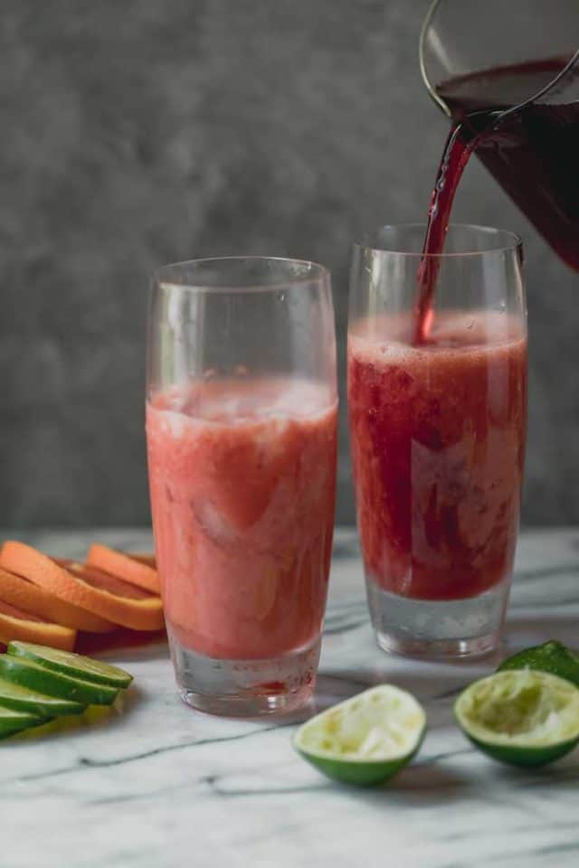 Dos vasos altos sobre una superficie de mármol con 3/4 de té helado de frambuesa llenos en un vaso y batido de té helado de frambuesa que se vierte en el segundo vaso.