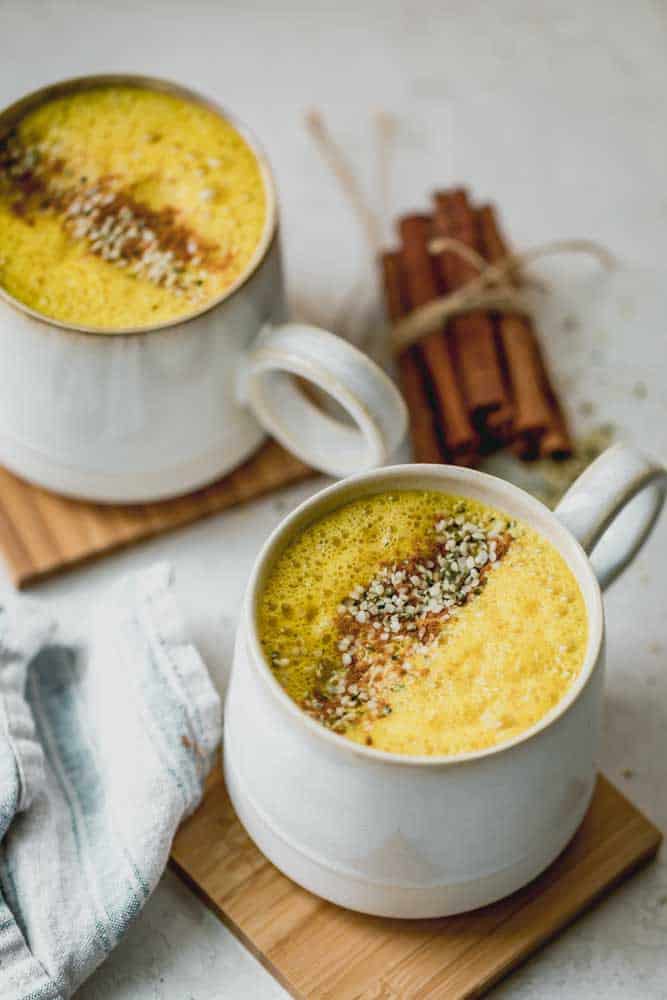 Dos tazas de café con leche de azafrán dorado con canela espolvoreada y semillas de cáñamo encima, junto con un montón de palitos de canela en el fondo