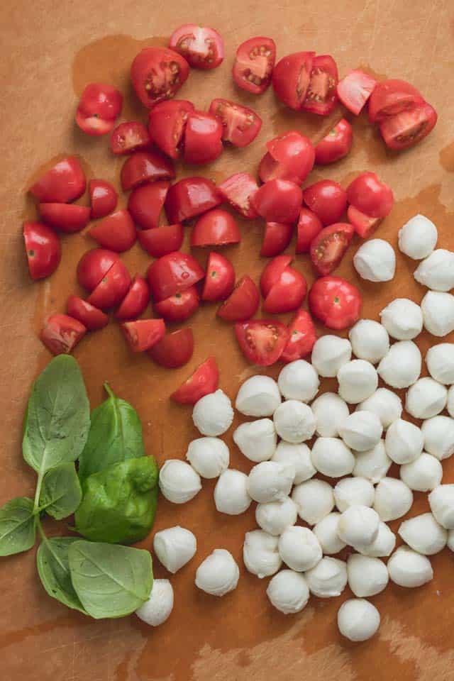 Tabla de cortar con pequeñas perlas de mozzarella, tomates cherry partidos por la mitad y hojas de albahaca fresca