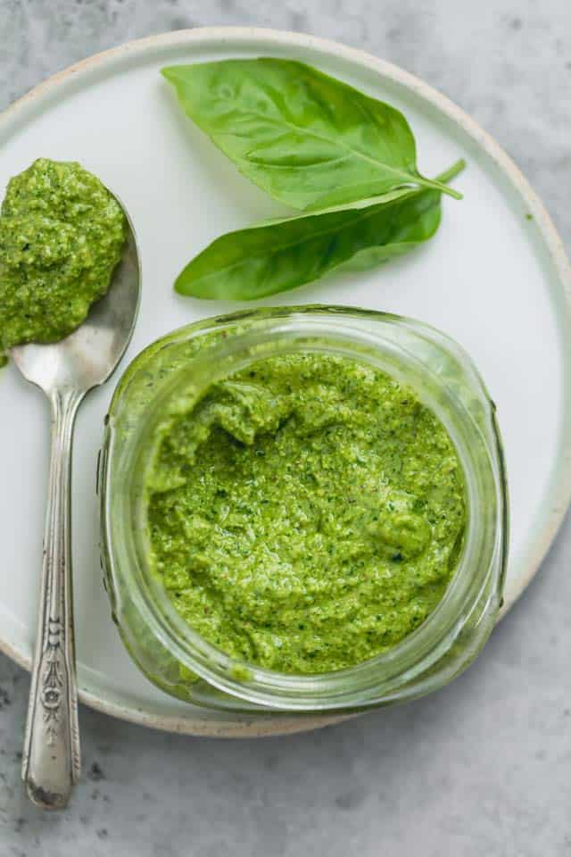 Frasco pequeño y ancho de pesto terminado con una cucharada de pesto al lado y un par de hojas de albahaca.