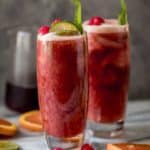 Dos vasos altos de batido de té helado de frambuesa con rodajas de naranja, gajos de limón, frambuesas frescas y hojas de menta adornadas en la bebida. Jarra de té helado o frambuesa en el fondo