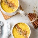 Dos tazas de café con leche de azafrán dorado con canela espolvoreada y semillas de cáñamo encima, junto con un montón de palitos de canela en el fondo
