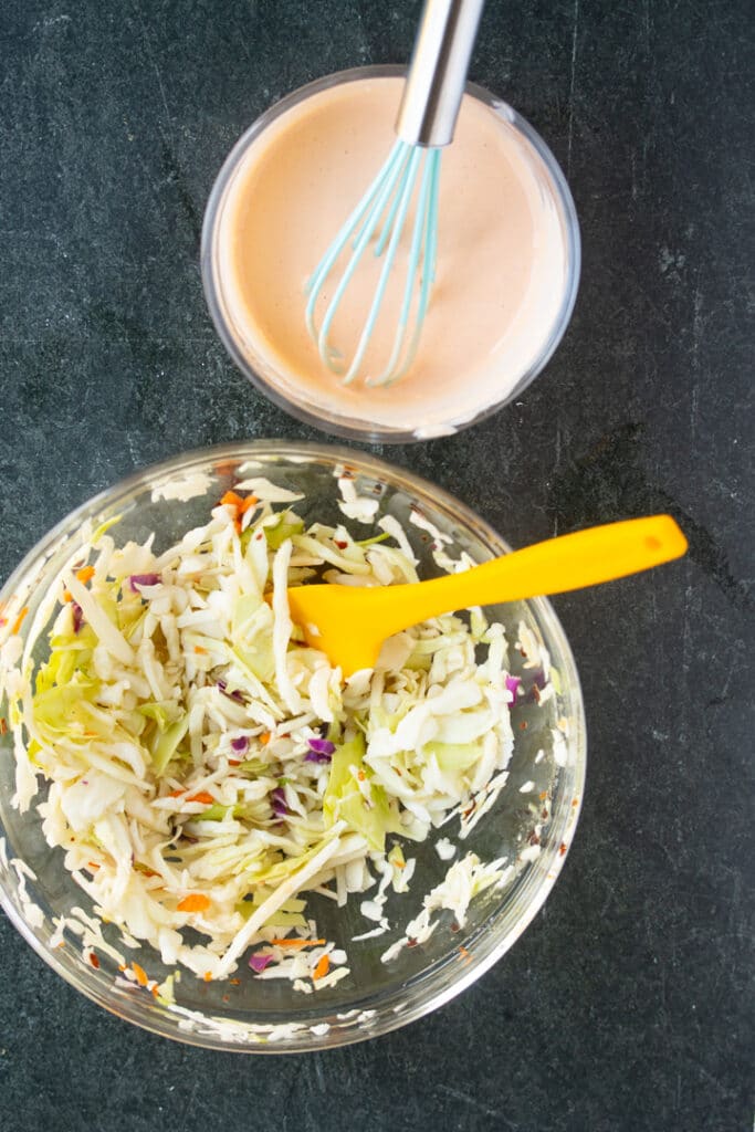 Mayonesa picante en un bol con un batidor que sobresale, ensalada de col en un bol con una espátula amarilla que sobresale